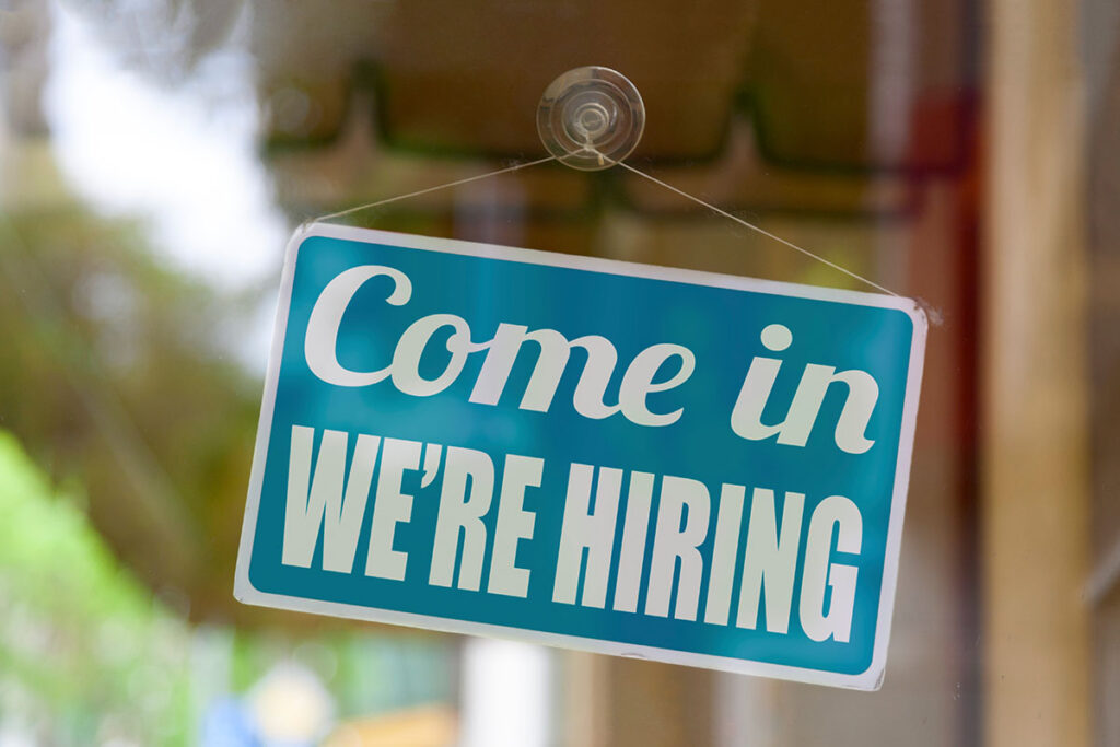 Close-up on a blue sign in the window of a shop displaying the message: Come in we're hiring.